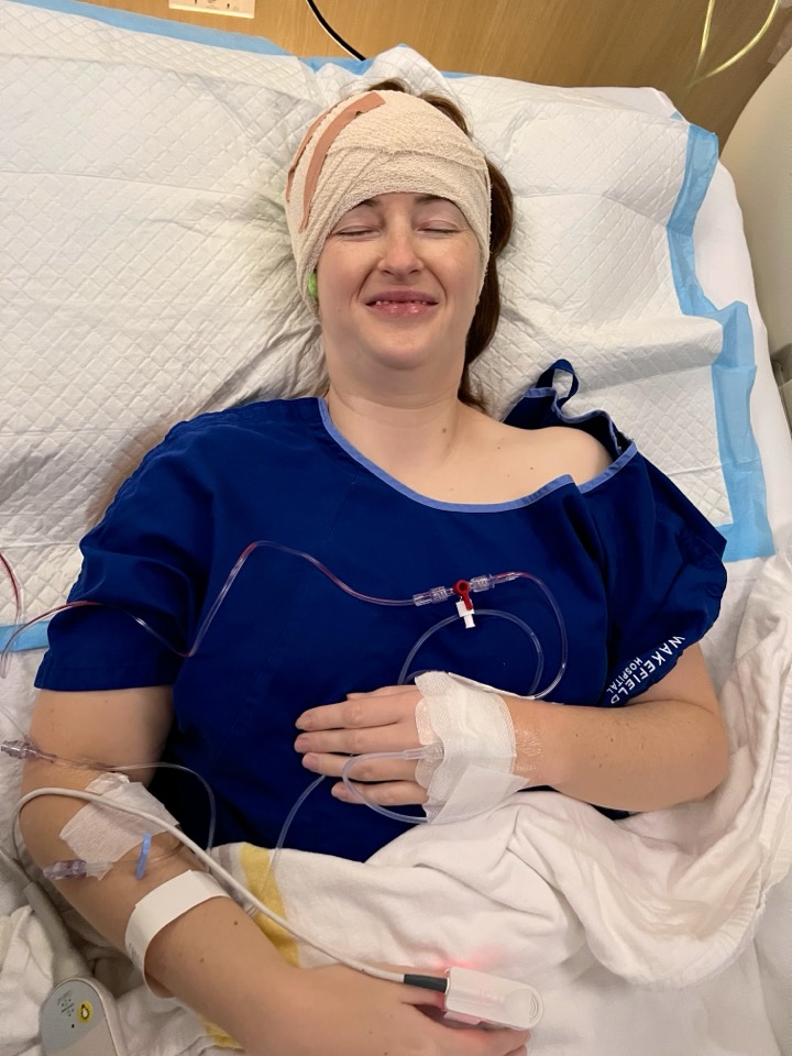 Heather grinning in her hospital bed right after surgery, head freshly bandaged