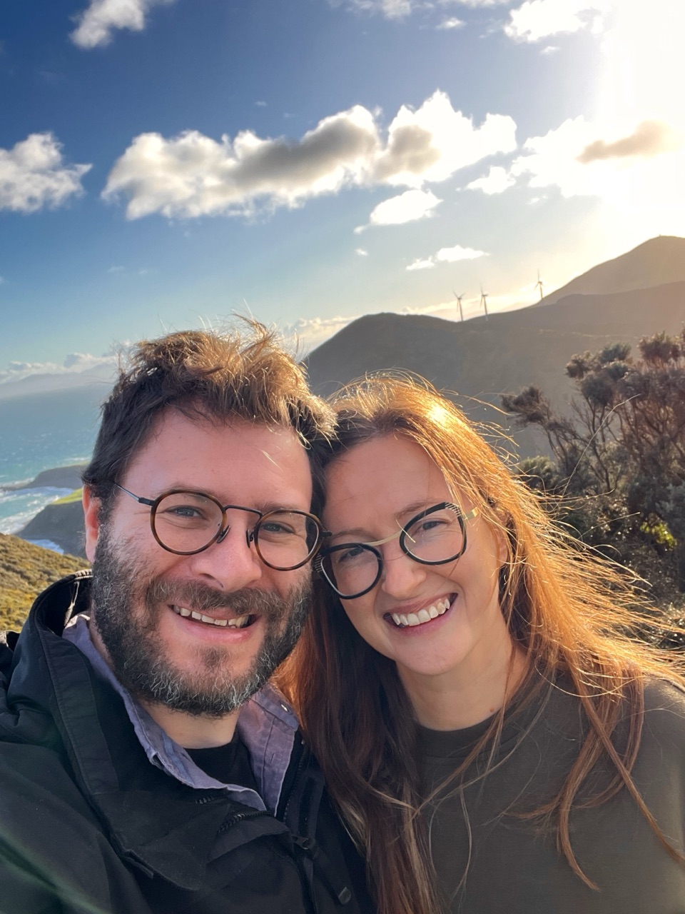 Heather and Kyle on a coastal hilltop, wind turbines behind