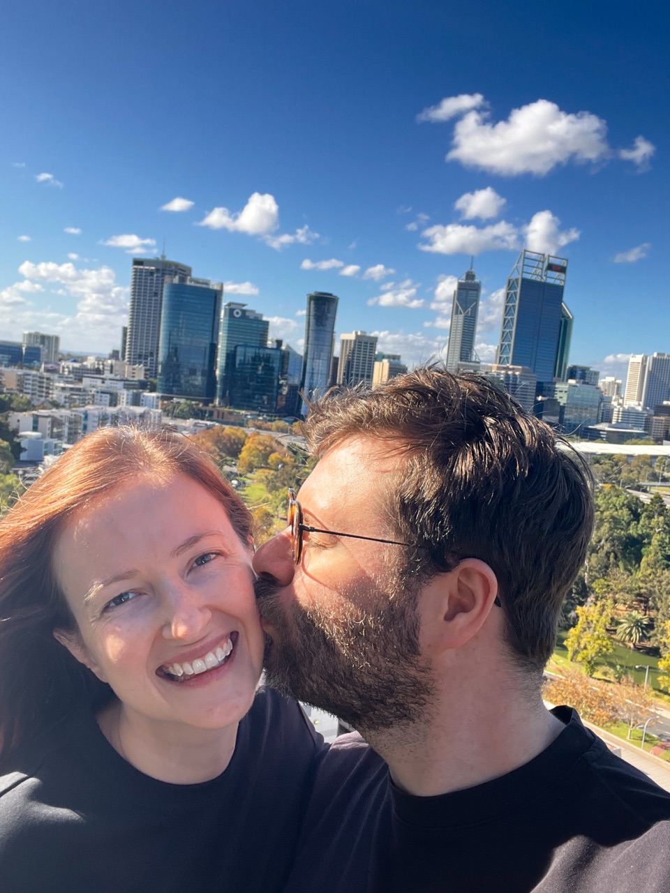 Kyle kissing Heather's cheek in front of a city skyline