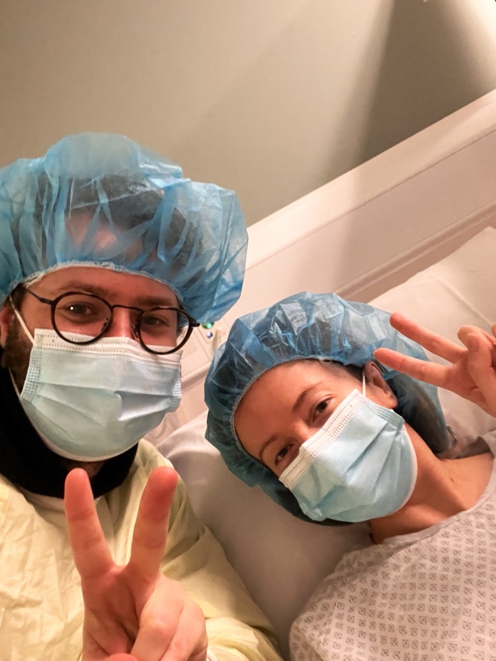 Heather and Kyle in surgical caps and masks, both flashing peace signs before surgery